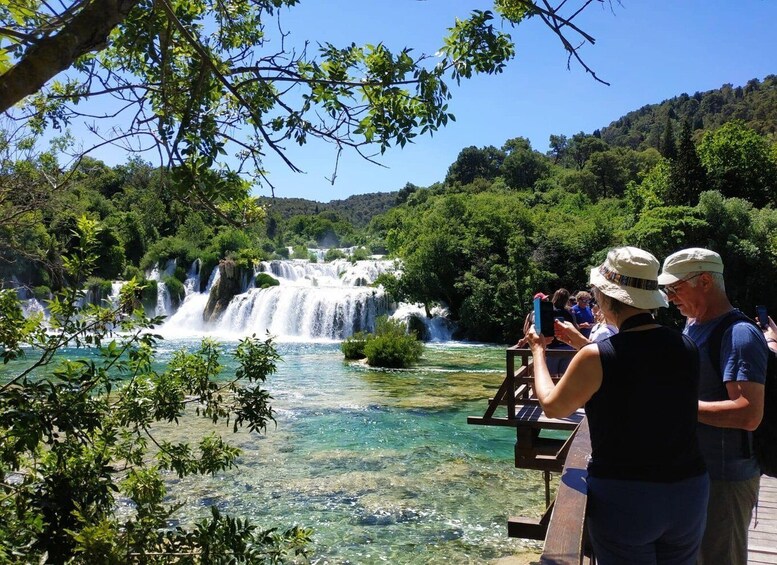A waterfall in a forested area with clear water and people taking photos. 1