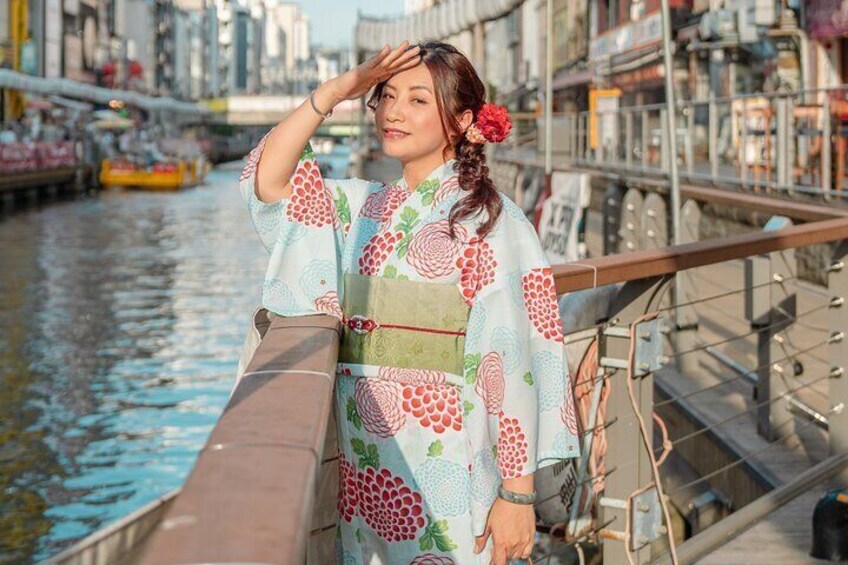 Een persoon in een kimono staat op een brug, met een rivier en gebouwen op de achtergrond. 1
