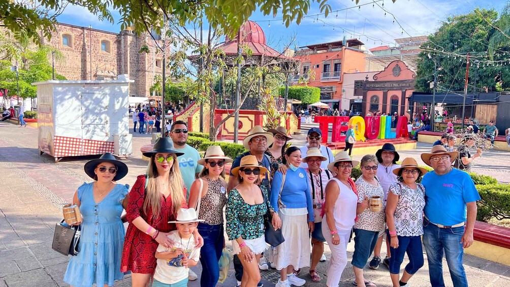 A group of people posing in front of a large Tequila sign in a plaza with historic buildings. 3