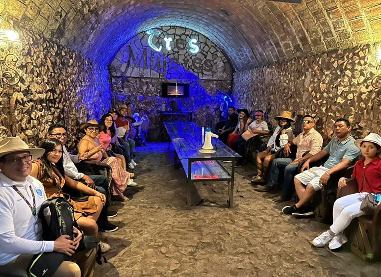 A group of people seated in a stone-walled room with a blue-lit archway and a table with a microphone. 2