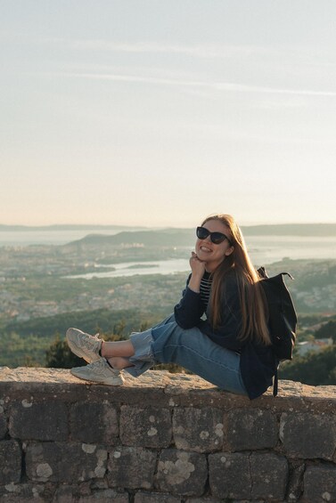 Una persona sentada en un muro de piedra mirando una vista urbana con un río y colinas. 3