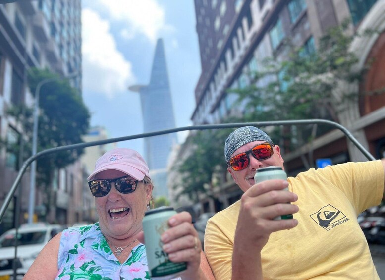 Two people in a convertible car, one holding a drink, with city buildings and a tall skyscraper in the background. 4