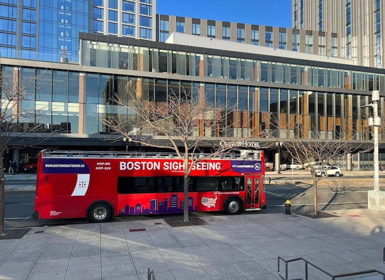 A red bus with 'BOSTON SIGHTSEEING' and 'WWW.BOSTONSIGHTSEEING.ORG' on its side parked in front of a modern building with glass windows. 4