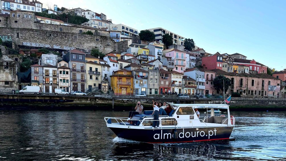 A boat named 'almdouro.pt' on a calm river with a backdrop of colorful buildings along the riverbank. 3