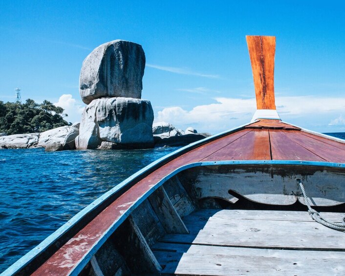 A wooden boat with a red and white hull, a blue railing, and an orange mast, anchored near a large rock formation in a body of water. 2