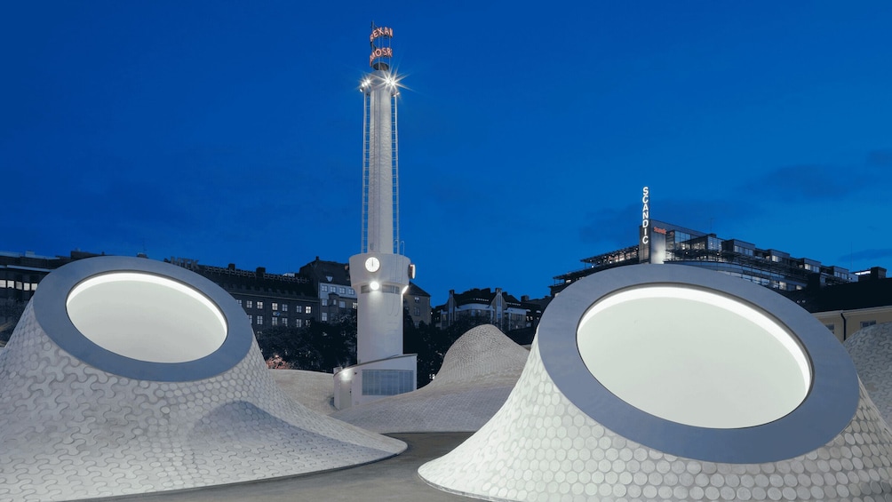 A modern sculpture with a tall, narrow tower and a circular base, set against a cityscape at dusk. 2