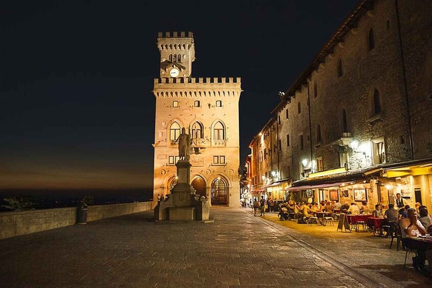 A historic square with a prominent tower and a statue in the center, surrounded by buildings with outdoor seating. 5