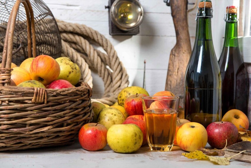A wicker basket filled with apples, a glass of amber liquid, and two green bottles. 2