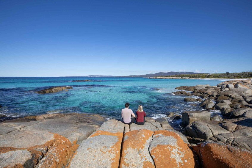Two people sitting on rocks overlooking a clear blue sea with a rocky shoreline and distant hills. 4