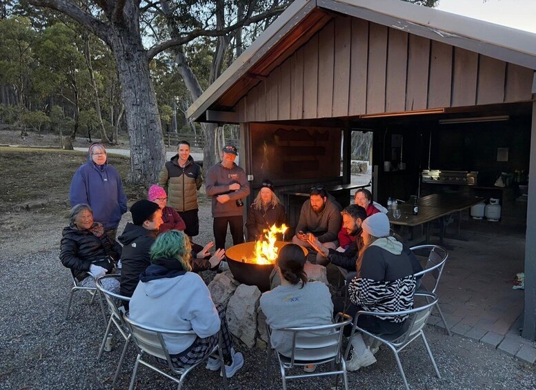 A group of people gathered around a fire pit in an outdoor setting with a wooden structure in the background. 2