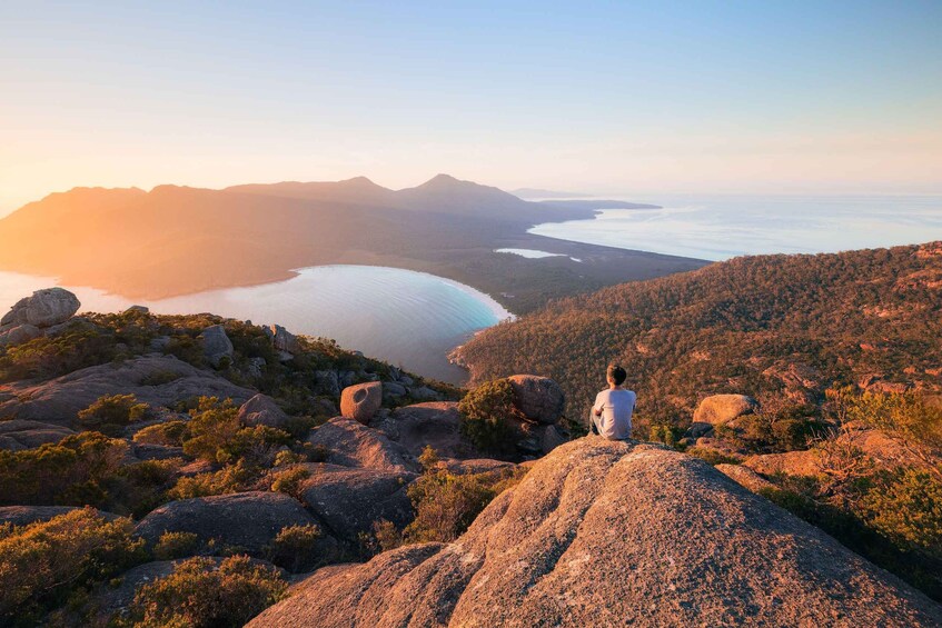 A person sitting on a rock overlooking a coastal landscape with mountains and a body of water. 1