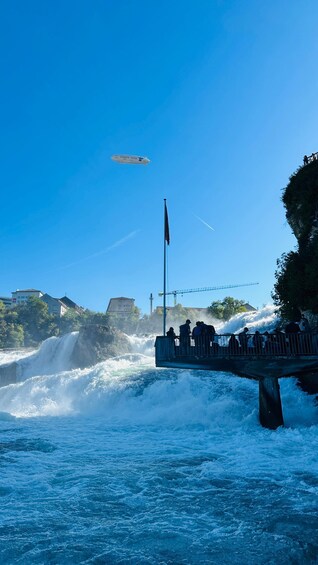 A river with a waterfall, a bridge with people on it, and a blimp in the sky. 1