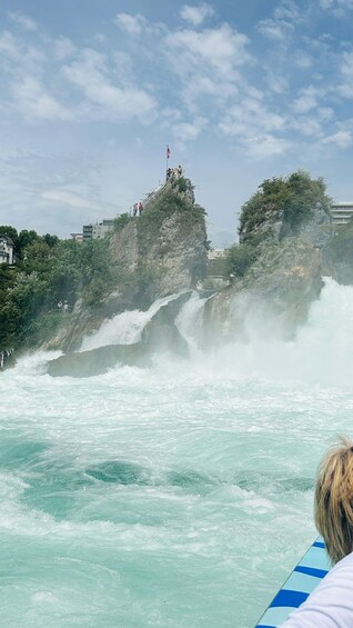 A waterfall cascading down a rocky cliff with a flag on top and people observing from a viewing platform. 2