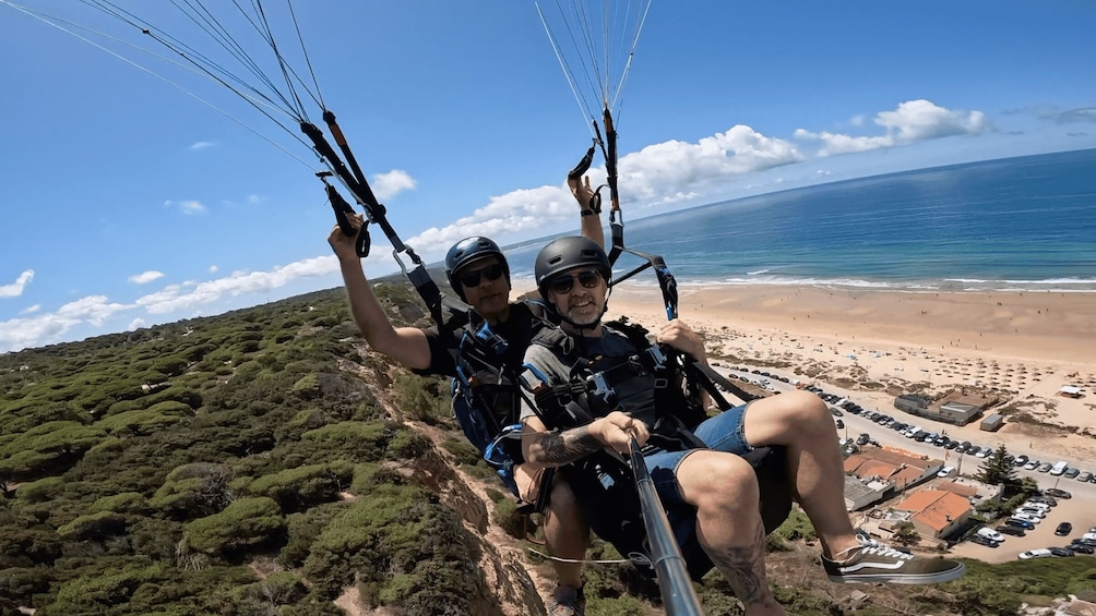 Two people paragliding over a coastal landscape with a beach and parked cars below. 2