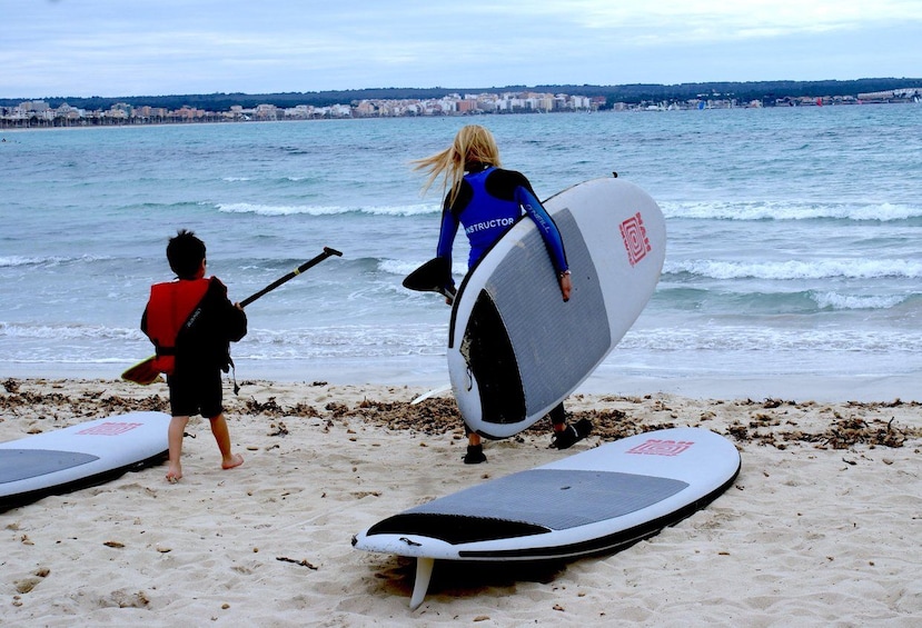 Una playa con dos tablas de paddle surf, una persona con traje de neopreno y un niño con chaleco salvavidas. 2