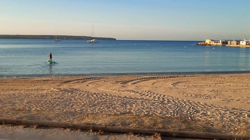 A sandy beach with footprints, calm water, a sailboat, and a distant shoreline. 2