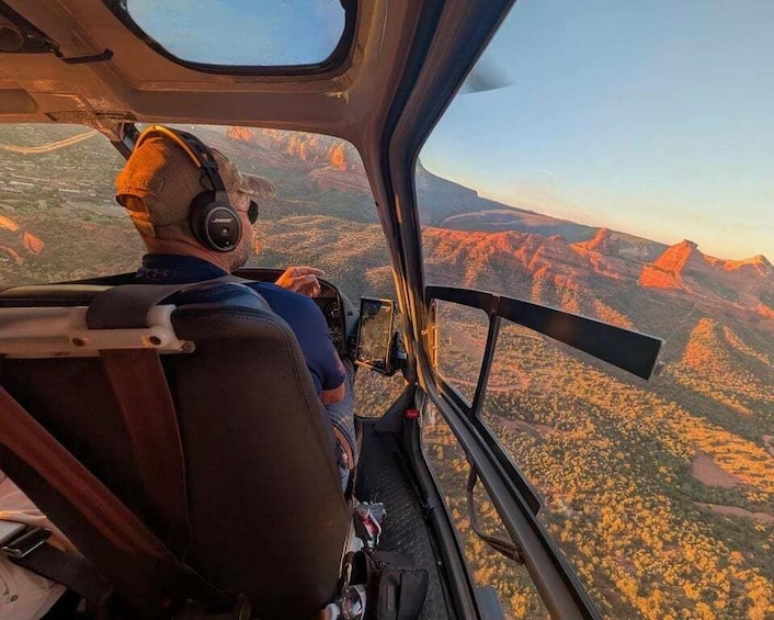 A helicopter pilot wearing a headset, with a mountainous landscape visible through the cockpit window. 1