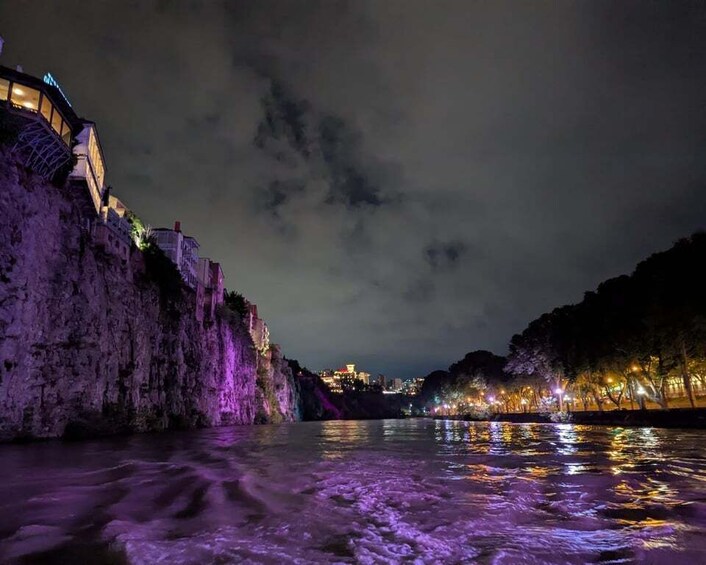 A nighttime river scene with illuminated buildings on a cliff and a cityscape in the distance. 2