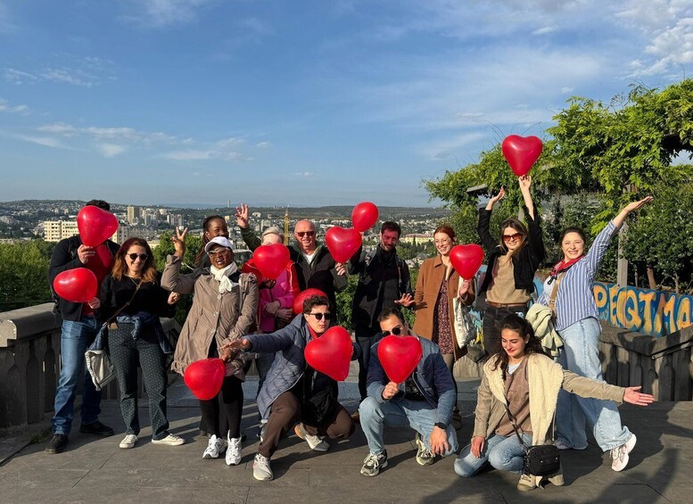 A group of people holding red heart-shaped balloons on a ledge with a cityscape in the background. 4