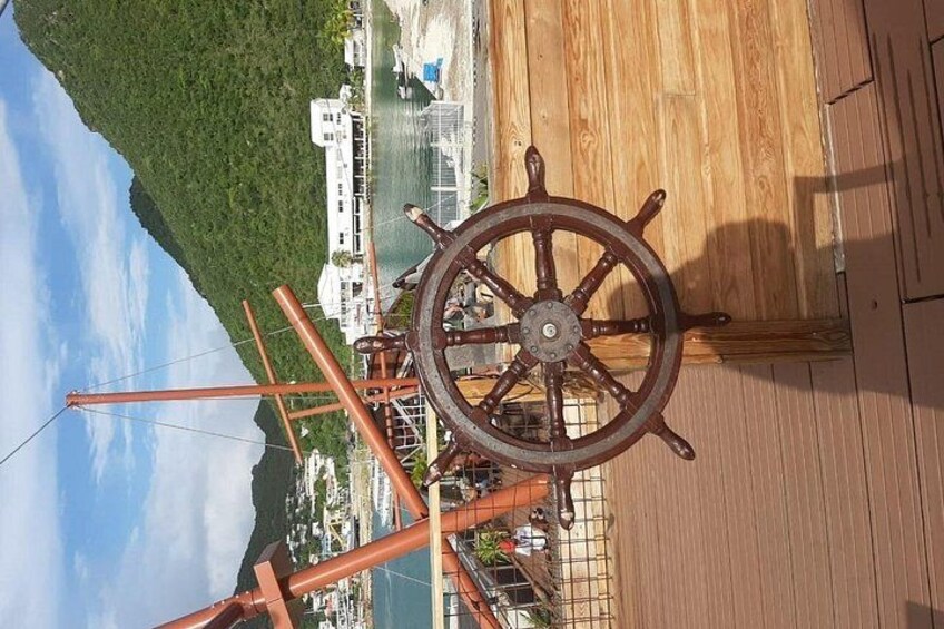 Cubierta de madera con timón, un muelle con barcos y un paisaje verde al fondo. 4