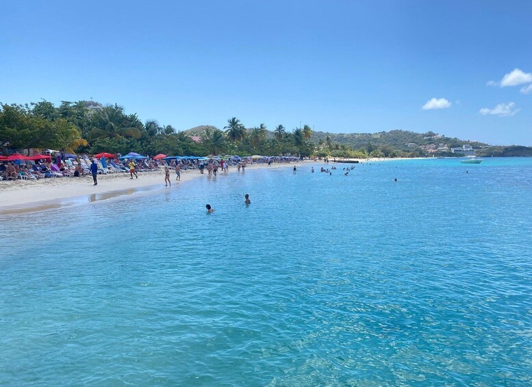 Een strand met mensen die zwemmen en zonnebaden, helderblauw water en een verre kustlijn met gebouwen. 2