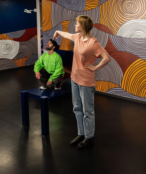 Una persona con remera color melocotón y jeans de pie junto a otra persona con remera verde, sobre un banco azul, con un llamativo mural abstracto de fondo. 5