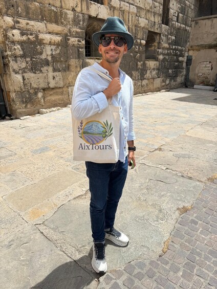 A person standing in front of a stone building with a vintage hat, a light blue shirt, and a tote bag with a logo. 2