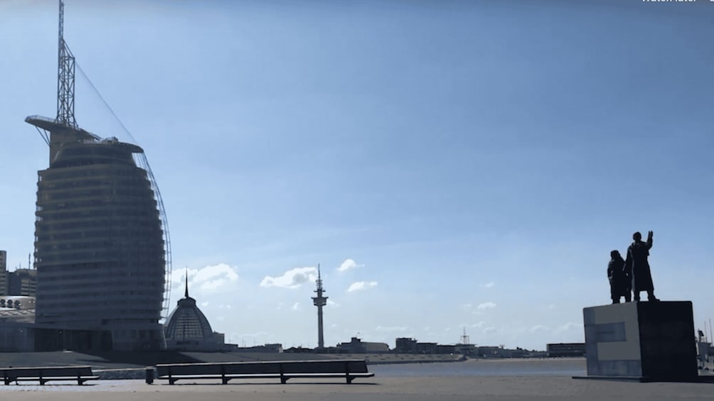 A coastal cityscape with a distinctive sail-shaped building, a lighthouse, and a statue of two figures on a pedestal. 3