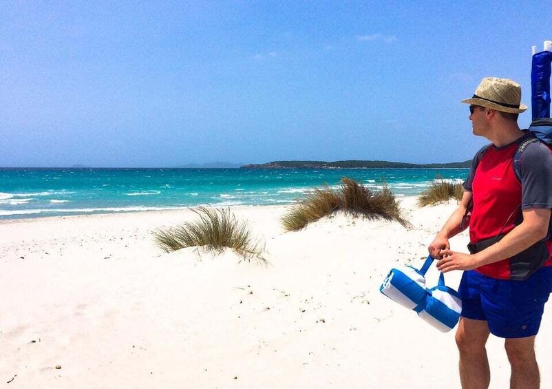 Una persona en la playa sosteniendo una bolsa azul y blanca, con agua azul cristalina y arena blanca al fondo. 2