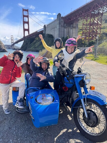 A group of people on a motorcycle with a blue sidecar, a bridge in the background, and a clear sky. 3