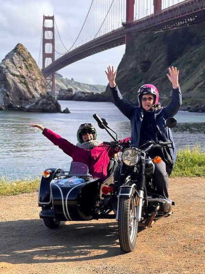 Two people on a motorcycle with a sidecar, waving by a bridge and water. 4