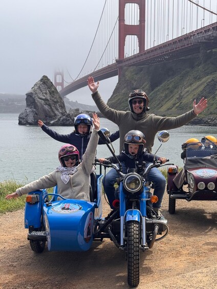 A group of people on a motorcycle with a sidecar, a child in a sidecar, and a vintage car parked near a bridge. 2