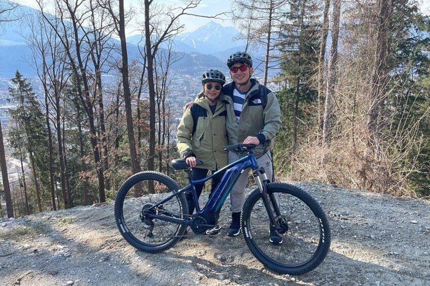 Due persone in piedi accanto a una mountain bike su un terreno roccioso, con alberi e montagne sullo sfondo. 3