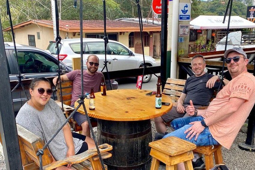 Four people seated at a wooden table outdoors with a beer bottle on it. 5