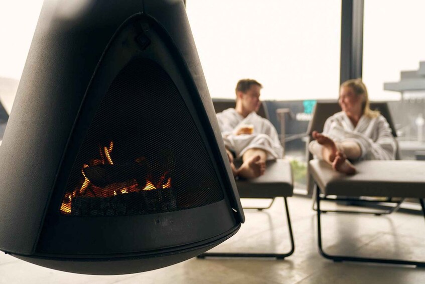 Una chimenea moderna con fuego visible, dos personas relajándose en sillones y una ventana grande al fondo. 3