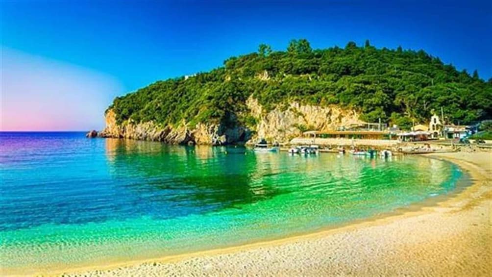 A beach with clear turquoise water, a sandy shore, and a forested hill in the background. 1