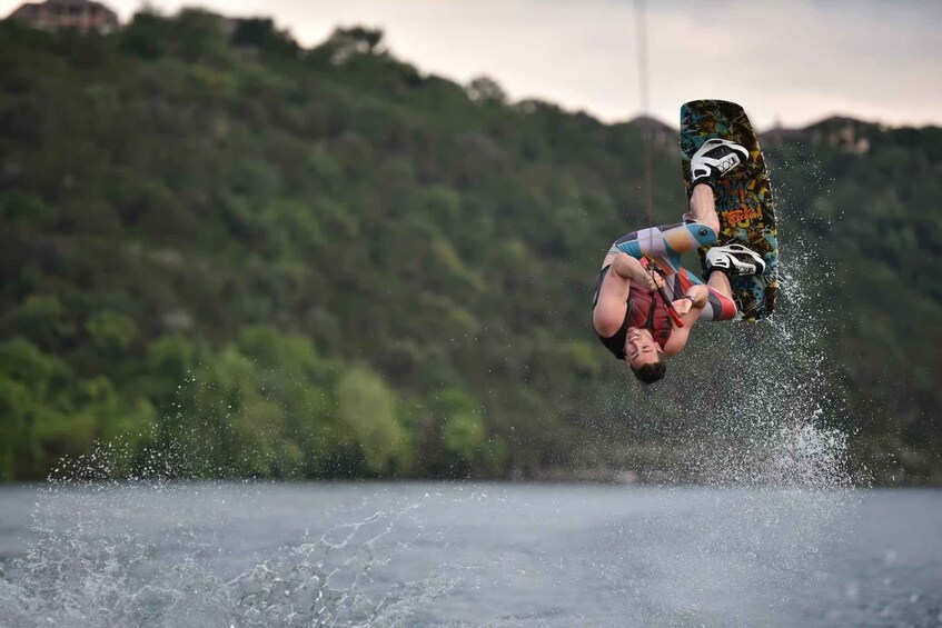 Una persona realiza un salto atrás con una tabla de wakeboard sobre el agua, con una colina boscosa al fondo. 3