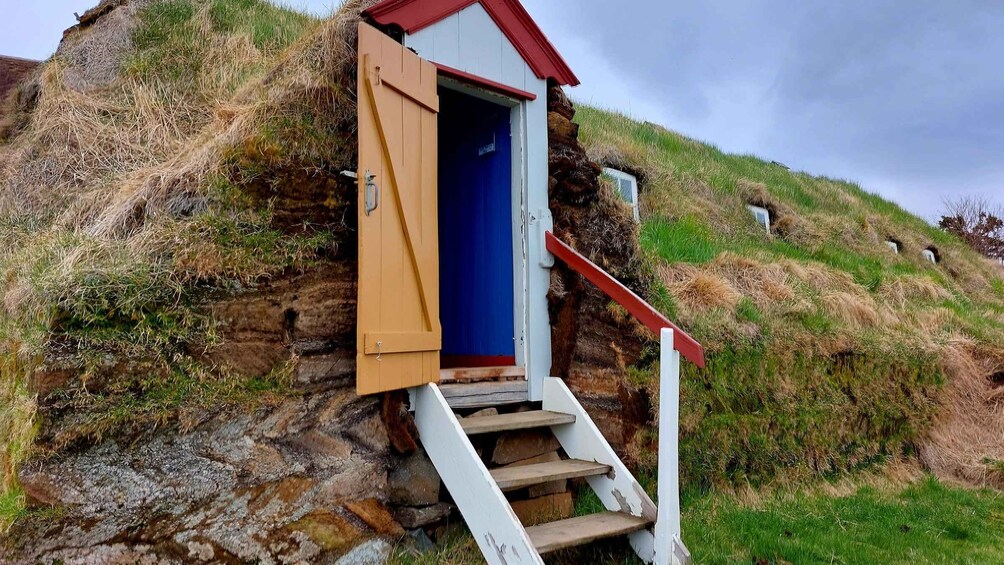A wooden structure with a red roof and a blue door, situated on a grassy hill with a stone wall. 2