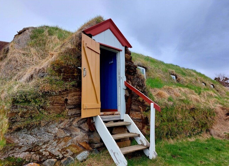 A small wooden structure with a red roof and a blue door, built into a grassy hillside. 2