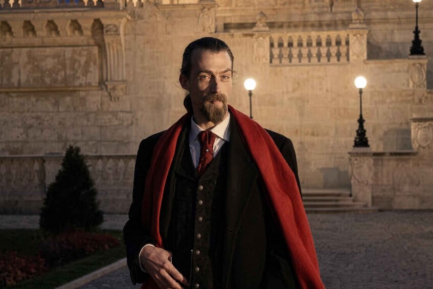 A man in a dark coat with a red cape standing in front of a stone building with arched windows and a statue. 1