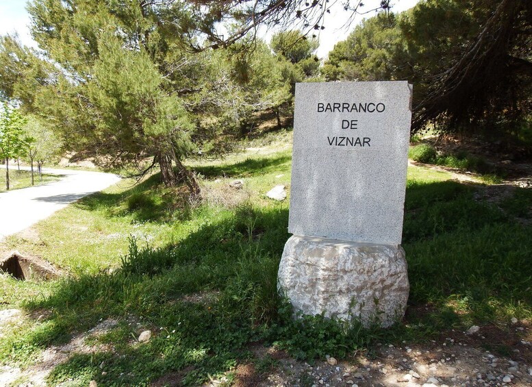 Ett stenmonument med inskriptionen ”BARRANCO DE VIZNAR”, placerat i en naturlig miljö med träd och en grusväg. 5