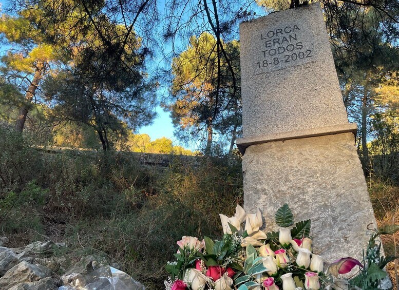 Ett stenmonument med inskriptioner, omgivet av träd och en blomsterarrangemang. 4