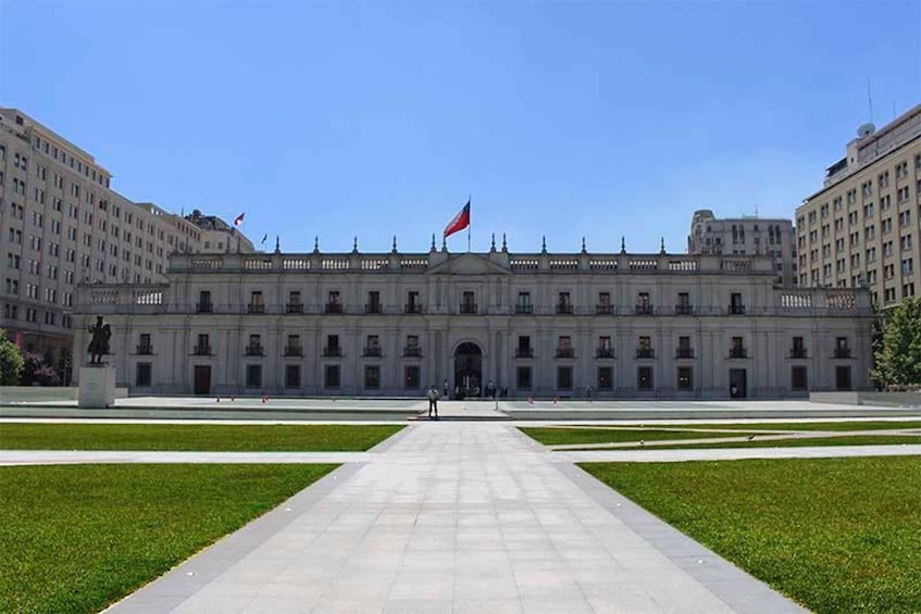 A large, historic building with a central entrance, flanked by two smaller structures, under a clear blue sky. 1