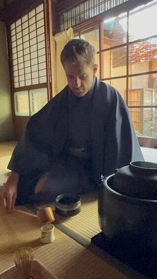 Une personne vêtue d’un costume traditionnel est assise à une table devant un grand pot et un petit bol, et semble préparer ou servir de la nourriture. 5