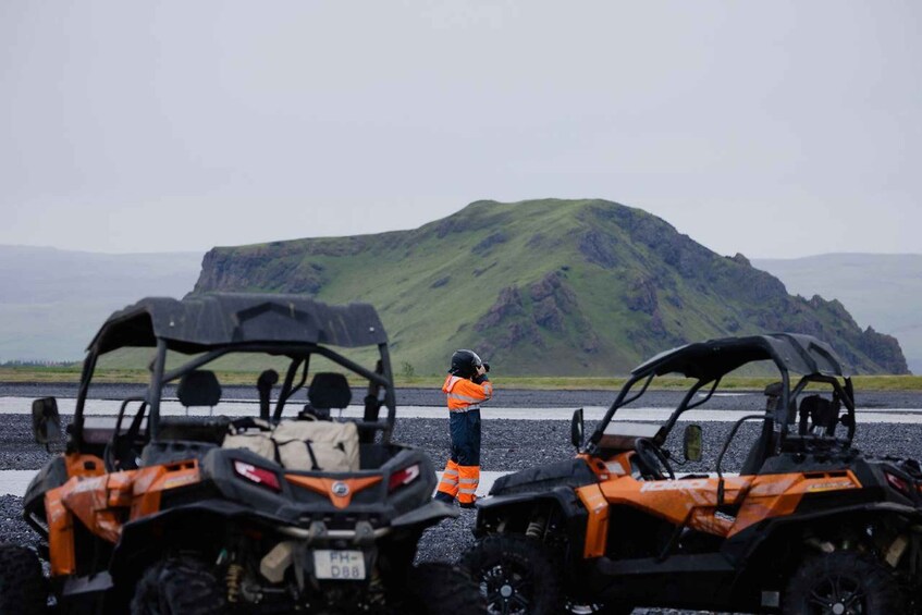 Two orange all-terrain vehicles parked on a gravel surface with a person in an orange vest standing between them. 2