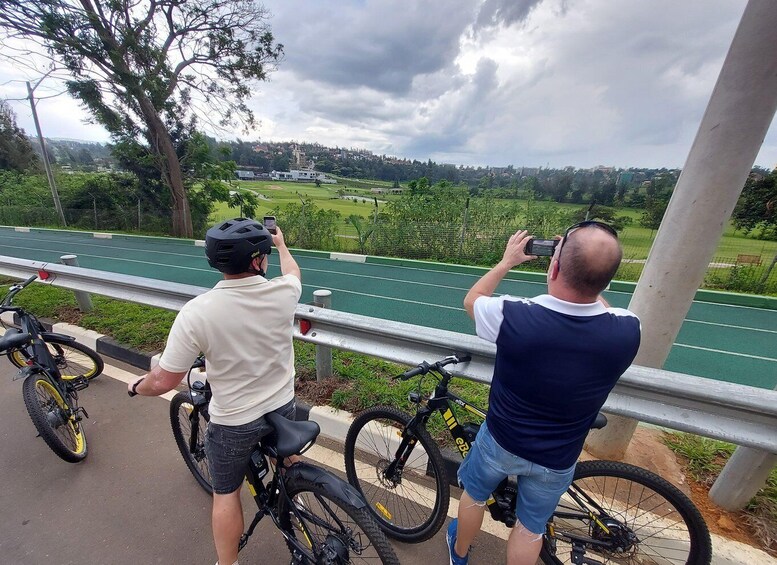Two people on bicycles, one taking a photo, overlooking a green track with trees and buildings in the background. 3