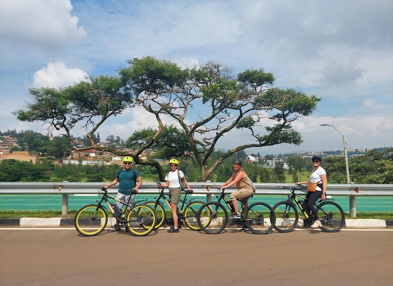 Four people on bicycles near a tree-lined bridge with a cityscape in the background. 1