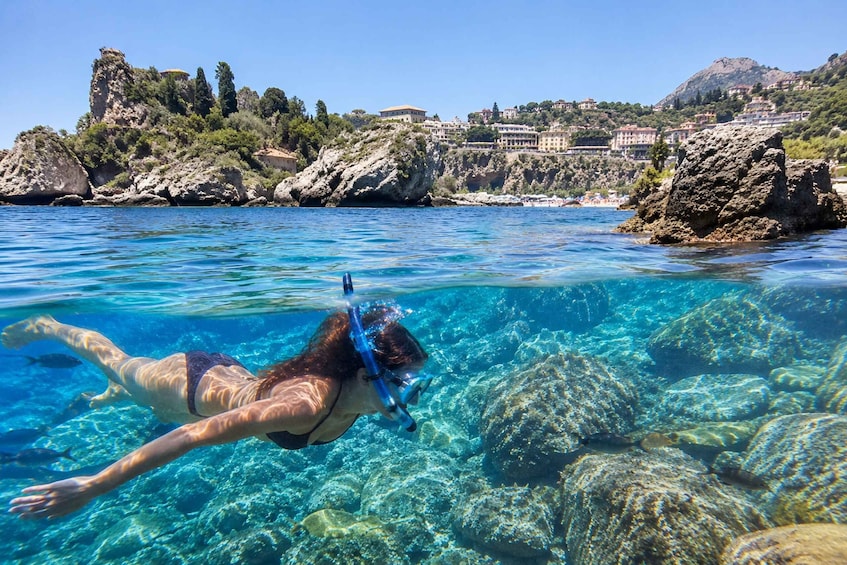 A person snorkeling in clear blue water with rocky formations and a coastal town in the background. 1
