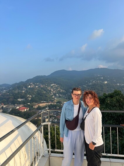 Two people standing on a balcony with a cityscape and mountains in the background. 5