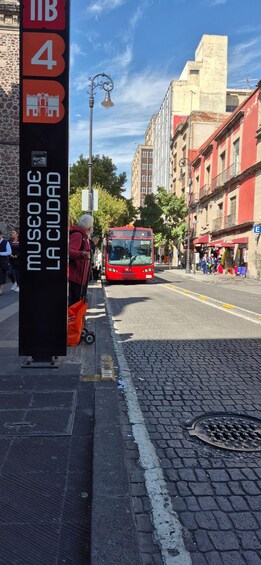 Une scène de rue avec un autobus rouge, une enseigne noire avec du texte blanc, et des bâtiments en arrière-plan. 2